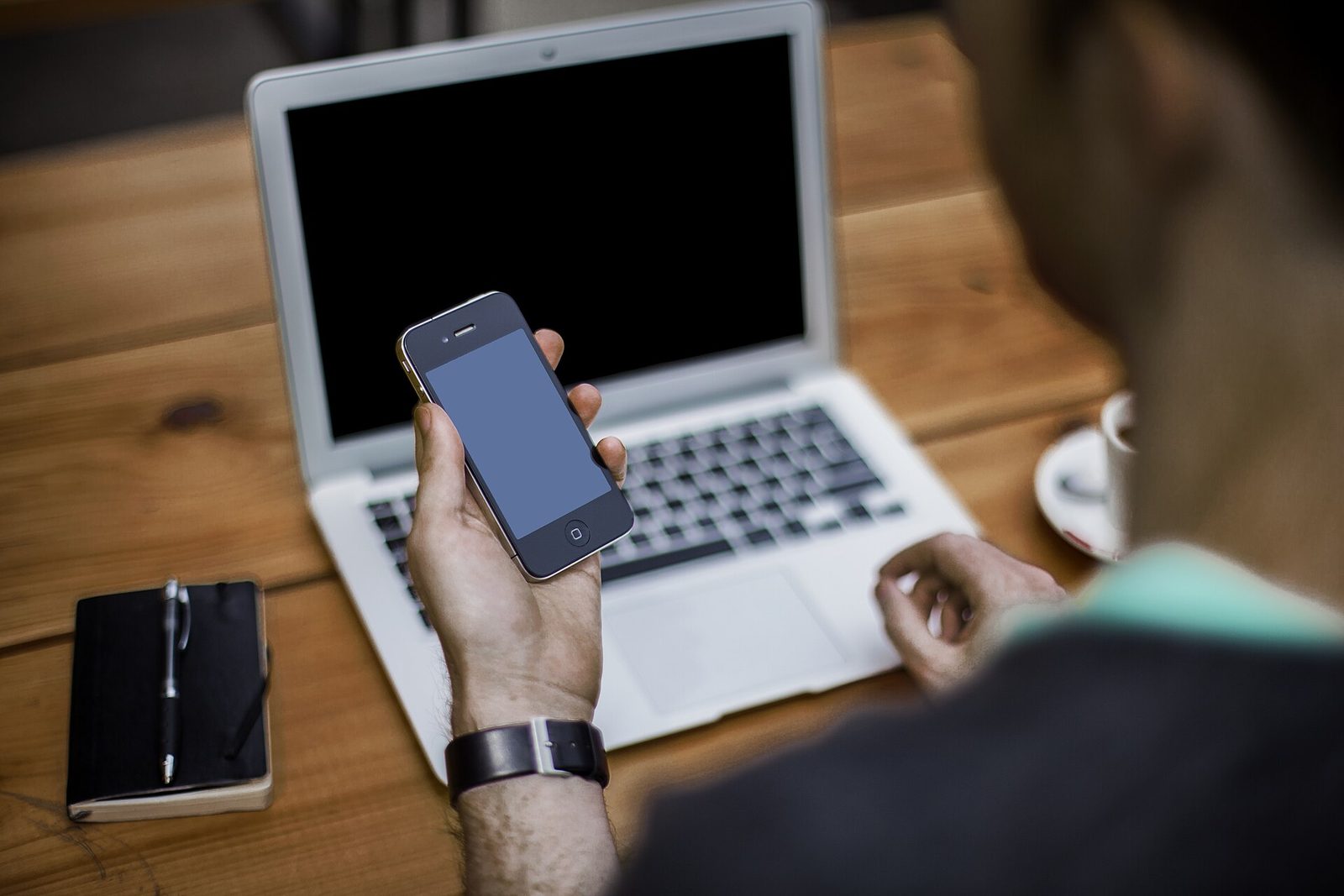 Homme assis à une table en bois, un smartphone dans une main, devant un ordinateur portable ouvert et un carnet noir